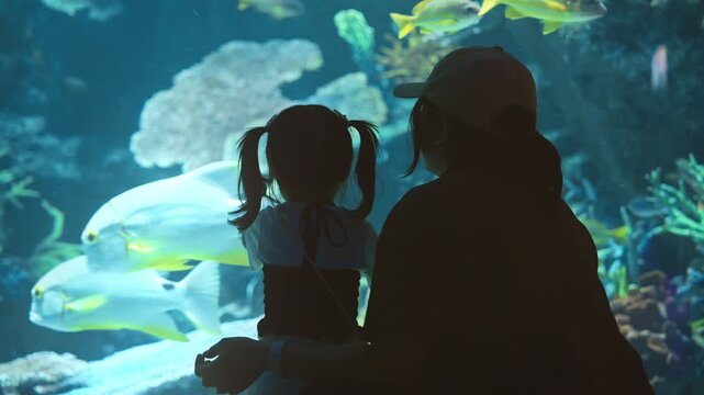Silhouette of mother and little daughter watching colorful tropical fish in large aquarium, family exploring underwater marine life together, childhood curiosity, bonding, education.
