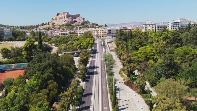 Aerial view flying backward along a modern tramway street revealing the ancient Acropolis of Athens, Greece, a famous landmark surrounded by the lush urban park and city buildings