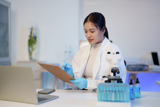 Asian scientist woman in lab coat and blue gloves researching, writing experiment data on clipboard in a modern laboratory for medical and scientific discovery