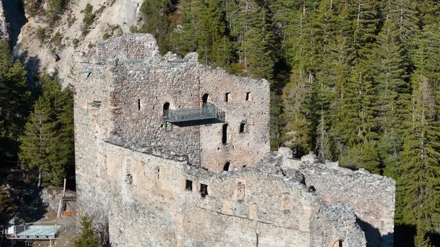 mountain road winds past belfort castle ruins rocky bluff pine forest aerial drone flyover arched window openings viewing platform railing graubunden switzerland surava albula valley heritage site