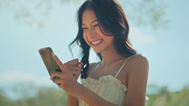 Excited young Asian woman looking at smartphone and raising fist in celebration outdoors, happy female reacting to good news, success, winning, achievement, mobile technology