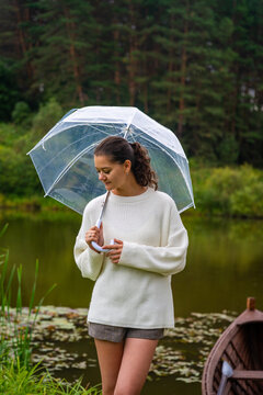 Young woman with curly hair holding a transparent umbrella stands by a serene pond surrounded by lush greenery and a wooden boat in the background