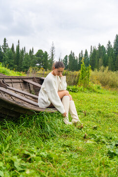 Young woman in white sweater and knee-high boots sits on an old wooden boat, surrounded by lush green grass and trees in a natural outdoor setting