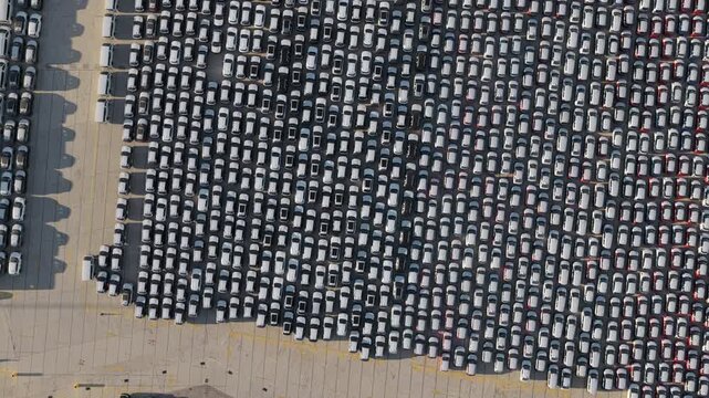 Countless new cars neatly parked in perfect rows at a huge automotive factory parking lot, awaiting distribution and export in a massive industrial port area, seen from a high drone