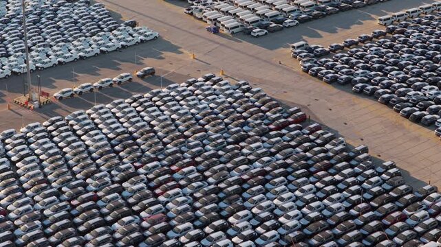 Thousands of new cars parked in perfect rows at a factory parking lot, awaiting distribution and export. Aerial perspective showing the vast scale of automotive manufacturing and global trade