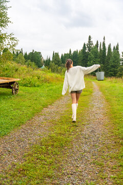 Young woman in white sweater and shorts walks along a gravel path carrying a bucket, with a wooden cart and trees in the background