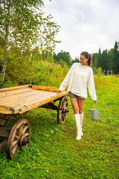 Young woman in white sweater and shorts walks beside wooden cart on grassy field, carrying a bucket, surrounded by trees and cloudy sky in rural landscape