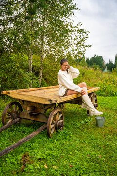 Young woman in white sweater and knee-high socks sitting on a wooden cart in a lush green field surrounded by trees and cloudy sky
