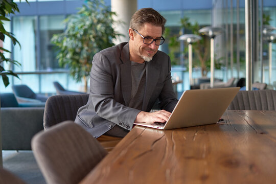 Happy middle age businessman using laptop computer in office. Older casual man working online on terrace of cafe, smiling.