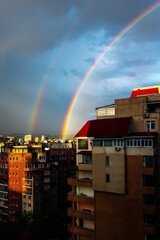 Obraz premium Vibrant double rainbow arching over residential cityscape during sunset with dramatic storm clouds. Urban landscape featuring bright sunlight illumination on building facades. Concept of hope, optimis