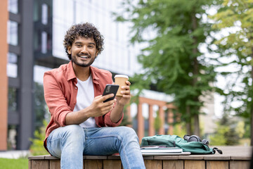 Fototapeta premium Happy college student holding takeaway coffee cup and smartphone outdoors resting between lectures.