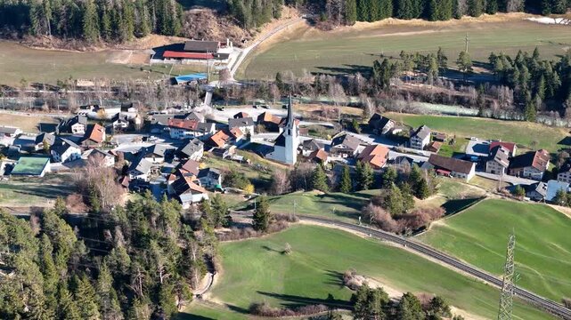 church tower crowns surava village rooftops chalets farmhouses cluster railway track winds through farmland forested slopes albula valley graubunden switzerland aerial drone footage close pass