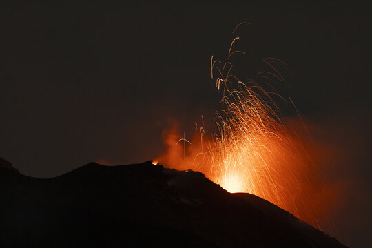 View of Stromboli volcano erupting at night with glowing lava and sparks shooting into the dark sky in Lipari, Sicilia, Italy.