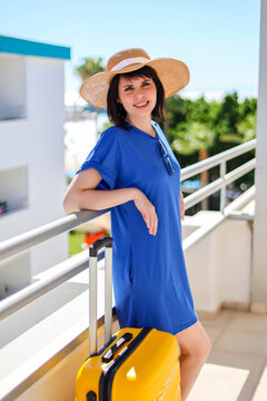 Happy woman tourist in blue dress and sun hat with bright yellow suitcase sunny hotel near her room enjoying summer vacation and travel.
