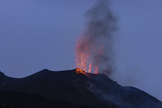 View of the Stromboli volcano erupting with glowing lava and sparks against a dark twilight sky in Lipari, Sicilia, Italy.