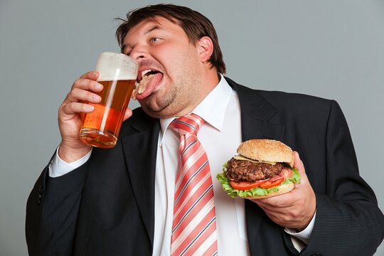 Overweight businessman eating hamburger and drinking beer in office environment with casual joy