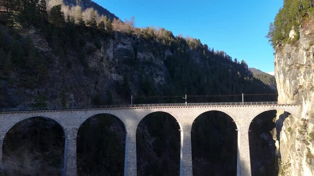 curved stone viaduct railway bridge crosses gorge near filisur graubunden switzerland limestone arches rise above forested slopes snowcapped alps aerial drone footage clear sunny mountain landscape