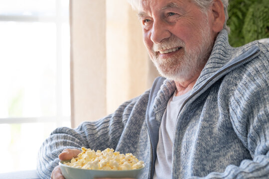 Senior man relaxing at home, enjoying a bowl of popcorn and smiling happily while watching tv, finding comfort in quiet leisure activities during retirement