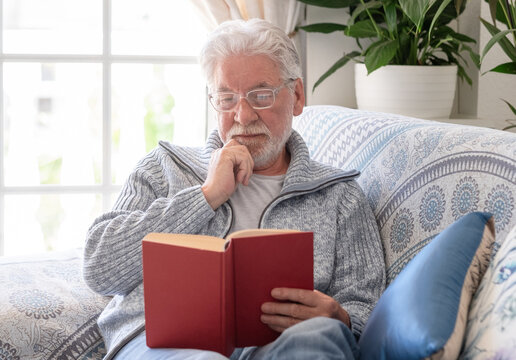 Mature adult man relaxing on a patterned sofa, reading a red book and wearing glasses, enjoying quiet leisure time at home during retirement, focusing on a hobby