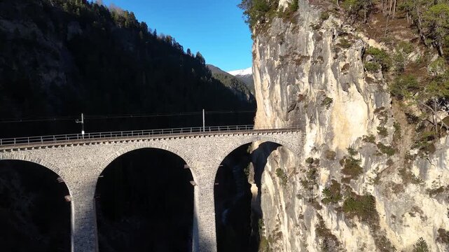 landwasser viaduct arches curve past towering limestone cliff near filisur switzerland sparse pine trees cling to rock ledges track runs along ravine edge aerial drone footage forested mountain gorge