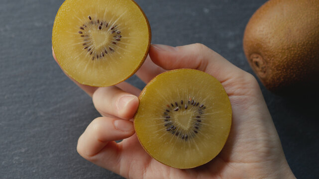 Close-up of a female hand holding two halves of a ripe golden kiwi, each showing heart-shaped core, on dark gray background with blurred whole kiwi for healthy eating, gourmet food, culinary content.