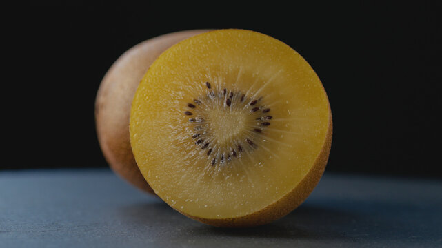 Macro shot of a ripe golden kiwi cut in half with a heart-shaped core, rich juicy pulp and seeds on a dark background with a blurred whole kiwi for healthy eating, gourmet food, and culinary content.