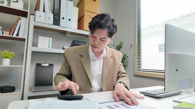 Asian male office worker using calculator for financial analysis in modern bright office