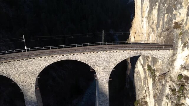 stone arches of landwasser viaduct curve beneath sheer limestone cliff near filisur switzerland shadow falls across rock pillars railway line spans forested ravine aerial drone footage alpine gorge