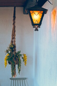 A warm glowing wrought-iron wall lantern mounted on a textured white wall with a soft blurred hanging macrame planter in the background