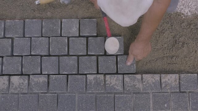 Top down view of a builder installing gray paving stones with a rubber mallet