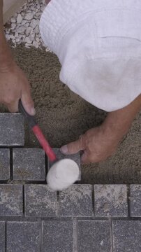 Vertical top view of a builder laying gray paving stones on sand with a rubber mallet