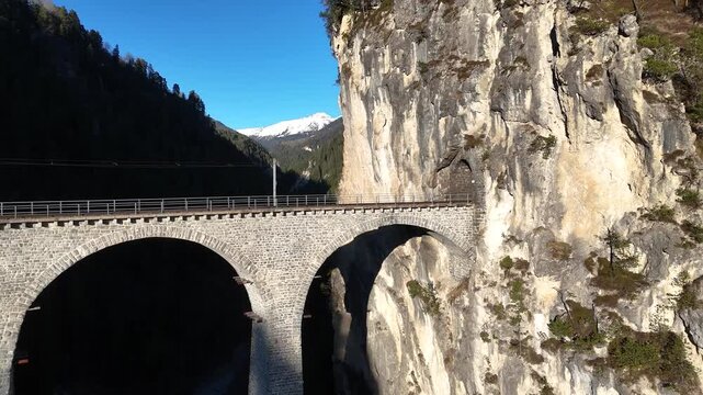 stone tunnel portal breaches limestone cliff landwasser viaduct arches curve below filisur graubunden switzerland snowcapped peak forested valley aerial drone footage clear blue sky mountain gorge