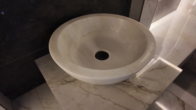 Matte beige round stone sink basin with central black slatted drain sits on polished marble countertop showing light tan and gray veining in modern bathroom setting, warm shadows and clean surfaces