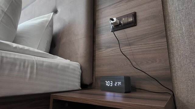 Neatly made bed in calm modern bedroom corner with striped white bedding, stacked pillows, wood headboard, bedside shelf, glowing digital clock, and wall outlet charger in soft warm light