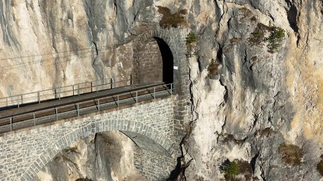 masonry tunnel archway carved weathered limestone cliff face landwasser viaduct track curves opening catenary wires vegetation rock ledges fissured aerial drone footage graubunden switzerland