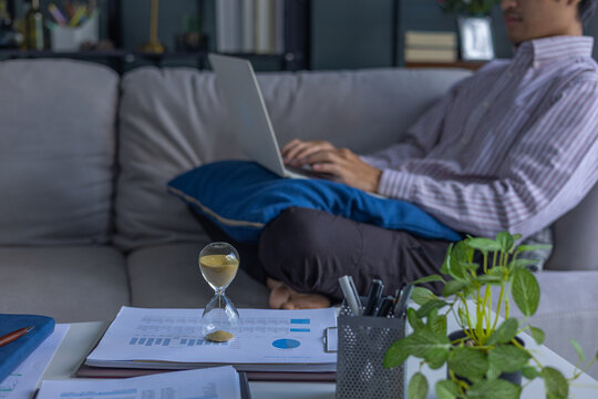 Man working on laptop sitting on sofa at home with running hourglass on the table. Concept for time management and extra over time working.