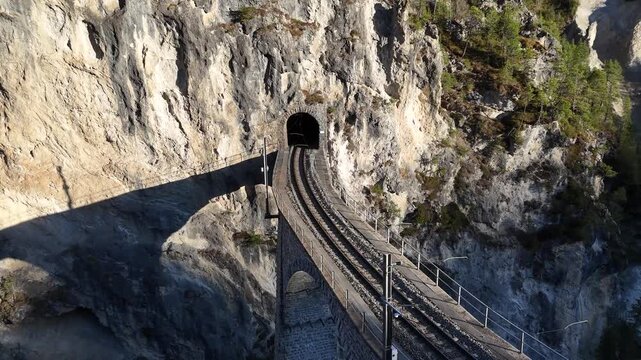 steel rails curve into landwasser tunnel entrance cut limestone cliff face shadow falls across viaduct arch below catenary pole lines aerial drone footage graubunden switzerland alpine gorge