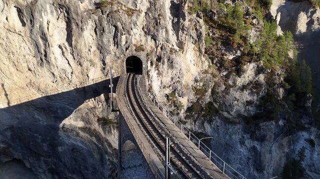 limestone cliff wraps around landwasser tunnel mouth curved track leads viaduct arches emerge below sparse pine trees cling rock ledges aerial drone footage clear sky graubunden switzerland alps