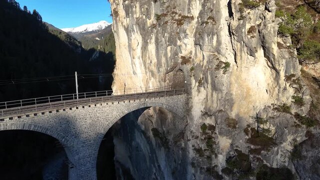pine tree frames landwasser viaduct curve tunnel portal limestone cliff filisur switzerland distant snowcapped peak forested valley catenary wires span track aerial drone footage clear sky graubunden