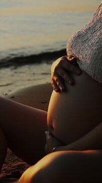 Close-up of pregnant woman gently touching and caressing her baby bump while sitting by the water at sunset. Tender maternity and prenatal bonding concept.