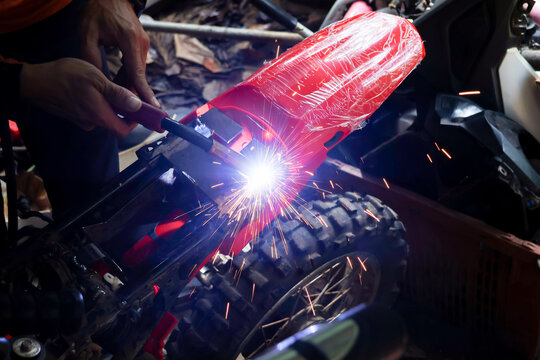 Welder repairing red motorcycle frame with bright sparks in workshop, close view showing skilled mechanical work and protective equipment around rugged wheel
