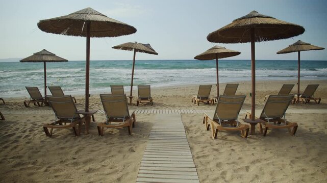 Empty sandy beach resort with wooden walkway, sunbeds and straw umbrellas near ocean waves