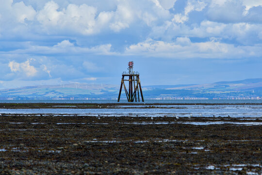 Navigation beacon on coastal mudflats at low tide with seabirds, distant hills and a dramatic partly cloudy sky.