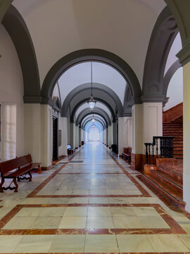 Long symmetrical interior corridor with repeated architectural arches, marble flooring, wooden benches, stairs and hanging lamps.
