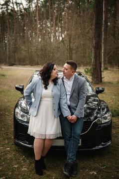 Loving man and woman standing and looking at each other near a black car parked in a pine forest.