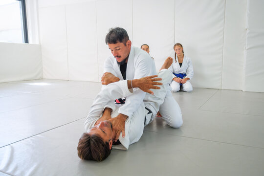 A Brazilian jiu jitsu black belt demonstrates an armbar setup from guard, controlling his partner's arm while positioning his legs as students observe