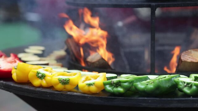 Colorful bell peppers and zucchini grilling on round barbecue grill with smoke. Fresh vegetables cooking over charcoal fire. Street food and outdoor barbecue concept.