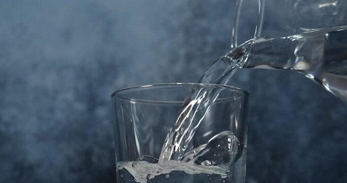 Water pouring from pitcher into glass with ice in slow motion
