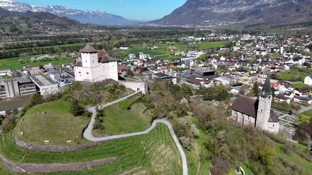 parish church clock tower cemetery grounds hilltop gutenberg castle round tower balzers town center modern buildings vineyard rows rhine valley snowcapped alpine peaks liechtenstein drone footage