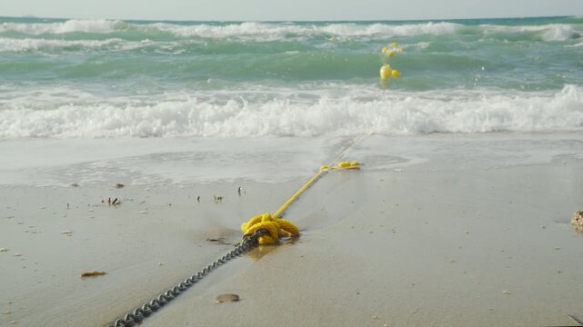 A yellow rope and metal chain lie on a sandy beach as rough ocean waves wash ashore towards floating yellow buoys